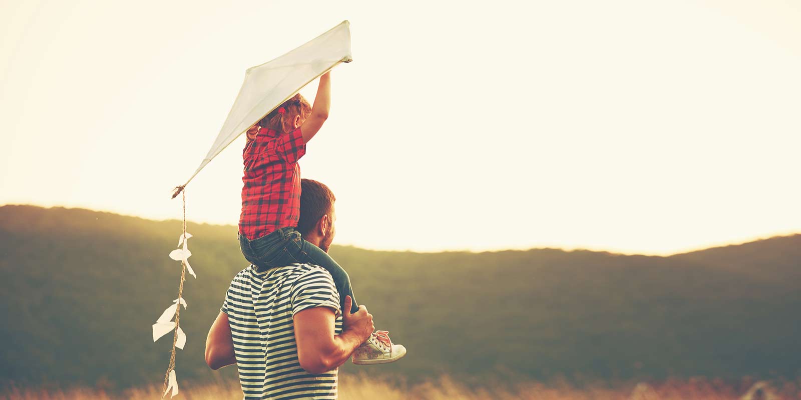 A kid holds a kite above his head as he sits on his father's shoulders.