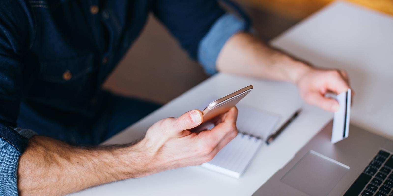 A man sits at a desk and holds a cellphone and a credit card. 