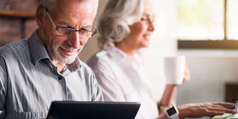 An elderly couple sits in their home as the man looks at his tablet. 