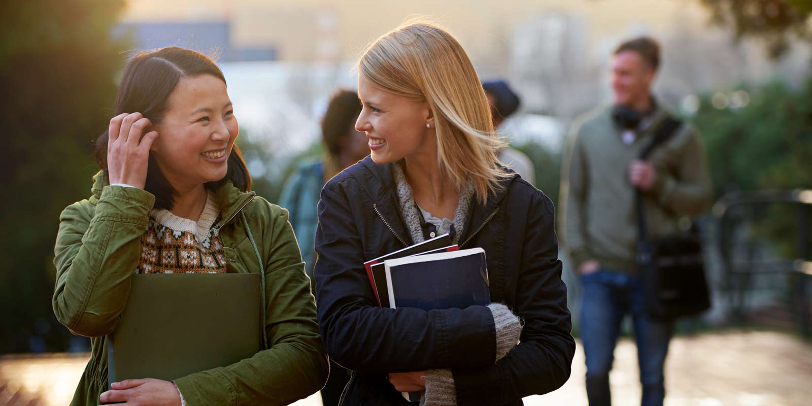 Two young women carry their notebooks and smile at each other as they walk outside.