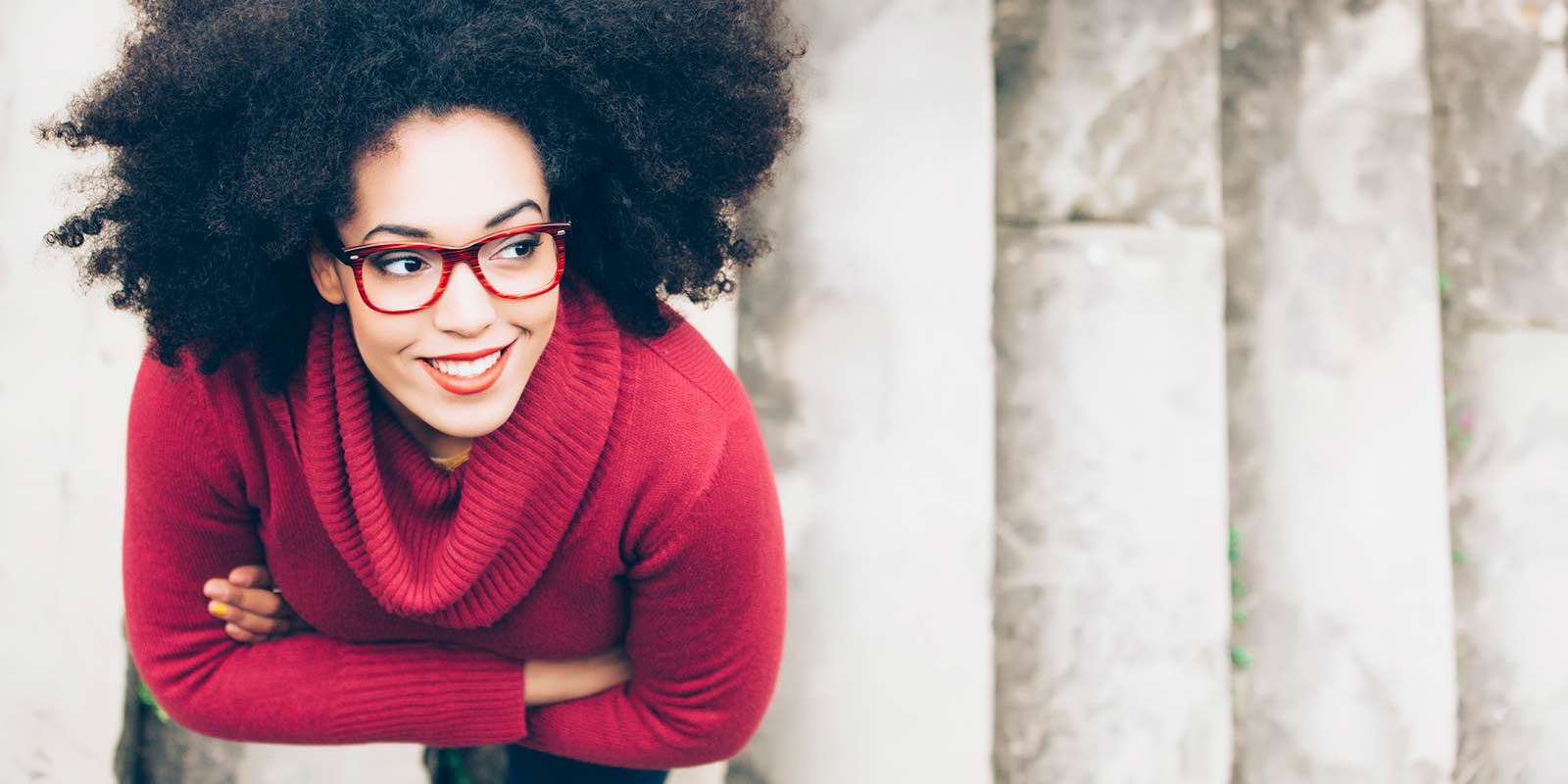 A young woman standing on concrete stairs looks up towards the sky and smiles.