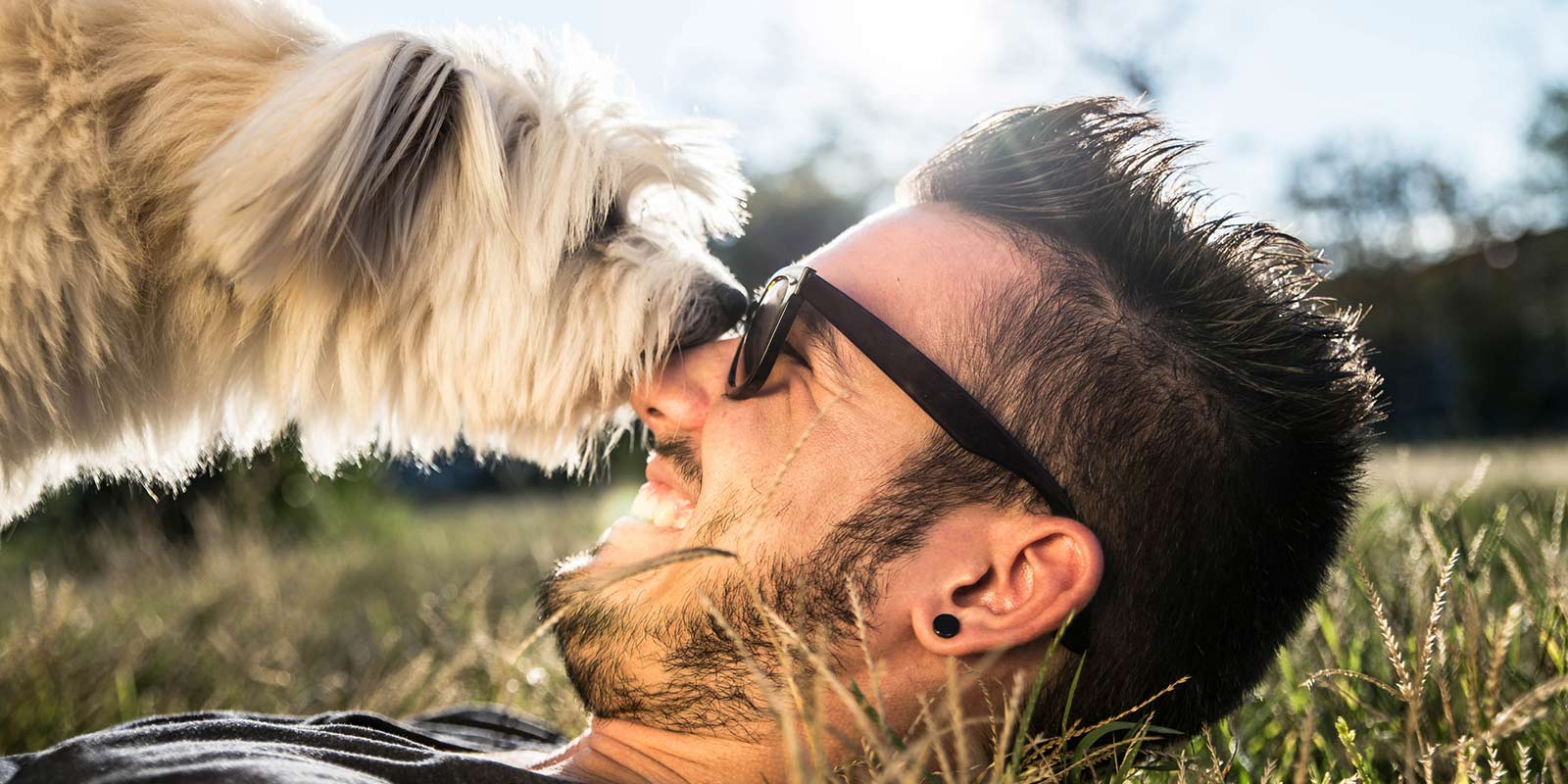 A man gets licked in the face by his dog as he is laying in the grass on a sunny day.
